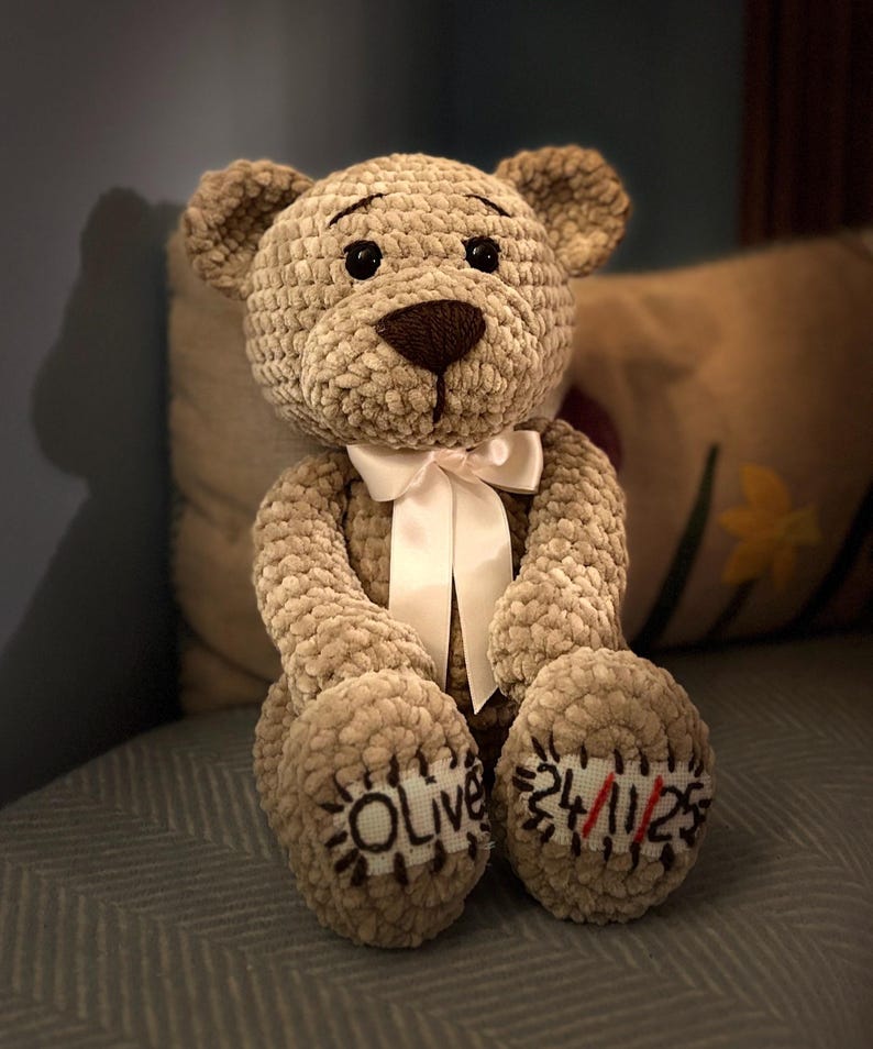 May include: A handmade, plush teddy bear in a soft, textured beige fabric. The bear has a brown snout and black eyes, with a white ribbon tied around its neck. The bear's paws are embroidered with the name "Olive" and the date "24/11/23".