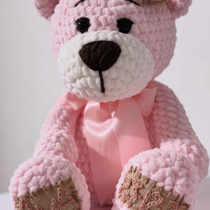 May include: A handmade pink crochet teddy bear with a white muzzle and a brown nose. The bear has black eyes, a pink satin bow, and patches on its paws. A small tag is attached to the ear.