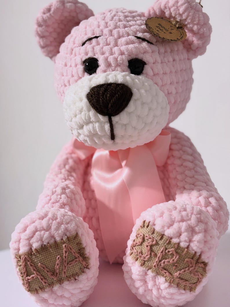 May include: A handmade pink crochet teddy bear with a white muzzle and a brown nose. The bear has black eyes, a pink satin bow, and patches on its paws. A small tag is attached to the ear.