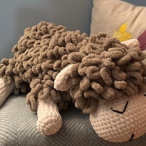 May include: A handmade, plush sheep pillow in shades of brown and cream. The sheep has a fluffy, textured body and a smiling face with stitched details. The pillow is designed to be soft and cuddly.