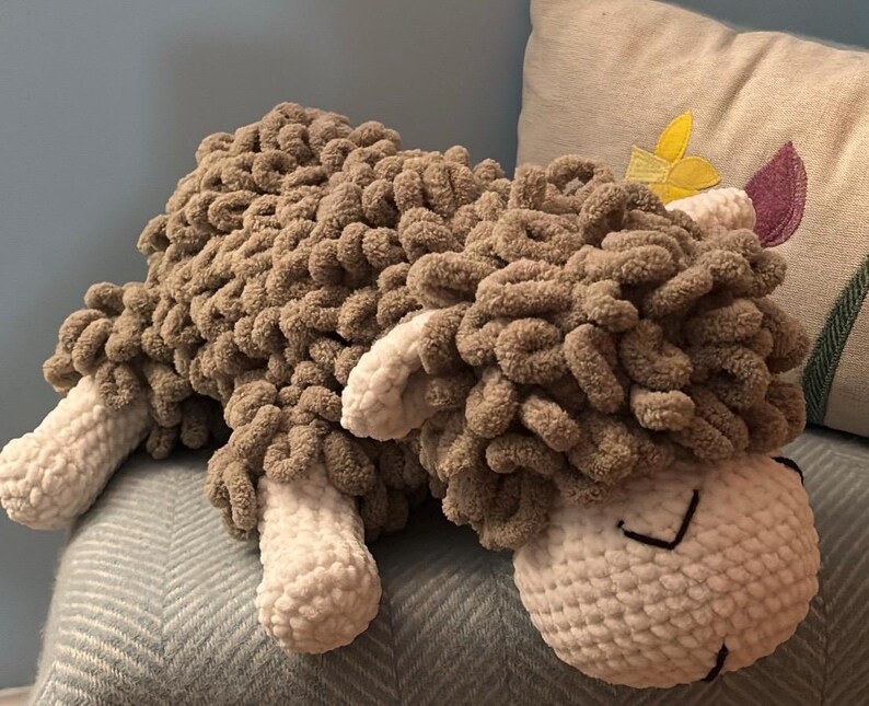 May include: A handmade, plush sheep pillow in shades of brown and cream. The sheep has a fluffy, textured body and a smiling face with stitched details. The pillow is designed to be soft and cuddly.