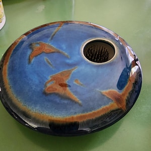 May include: A round, glazed ceramic ikebana flower vase featuring a deep blue center with brown, rust-colored accents along the edges. The vase has a circular opening off-center containing a metal kenzan pin frog for arranging floral stems.
