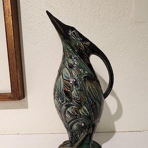 May include: A green and brown ceramic pitcher shaped like a bird with a long neck and a detailed design. The pitcher has a handle and a base with three toes.