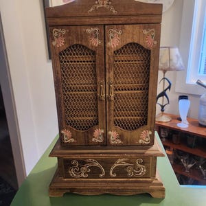 May include: A vintage wooden jewelry armoire with two doors and a base. The armoire is brown with decorative pink and gold accents. The doors have a wire mesh design. The armoire is sitting on a green surface.