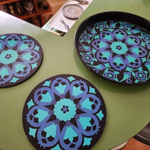 May include: A set of three round black trays featuring a repeating floral mandala pattern in shades of bright turquoise and deep blue. The circular trays have a smooth finish and a consistent geometric design across all three pieces.