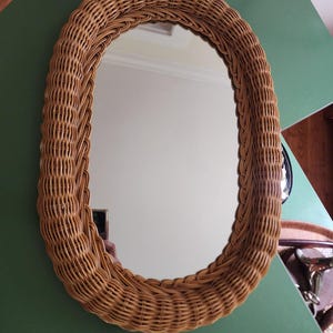 May include: Oval-shaped mirror with a woven rattan frame. The frame is a light brown color and has a textured surface. The mirror reflects the surrounding environment. The mirror is a decorative item.