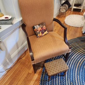 May include: A high-backed upholstered armchair with a small decorative pillow and a matching footstool. The chair has a light brown fabric with dark wood arms and legs. The footstool has a patterned fabric. A blue and white braided rug is beneath the chair.