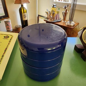 May include: A cylindrical, deep blue, multi-tiered stackable container with a smooth, glossy finish. The object consists of four distinct, rounded sections stacked vertically, creating a uniform, solid-colored, circular storage unit.