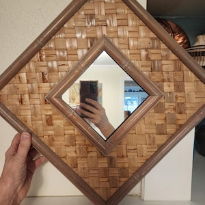 May include: Diamond-shaped wall mirror with a woven, light brown texture. The mirror is framed with a darker brown bamboo-style border. The inner mirror is also diamond-shaped, reflecting the surroundings.