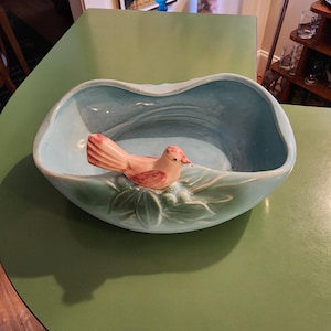 May include: A light blue ceramic bowl with an irregular, wavy rim. A sculpted pink bird with red detailing is perched on a cluster of green leaves and berries on the front exterior of the bowl. The interior of the bowl is a smooth, light blue.