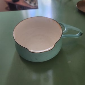 May include: A light blue enamel saucepan with a single handle. The interior is white, and the rim is outlined in black. The saucepan is sitting on a green surface.
