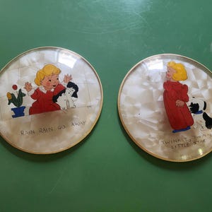 May include: Two vintage decorative plates with gold rims. Each plate features a cartoon illustration of a child in a red dress with a dog. One plate has the text "Rain Rain Go Away", the other reads "Twinkle Twinkle Little Star".