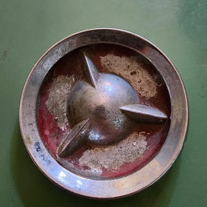 May include: A circular vintage automotive hubcap featuring a polished chrome outer ring and a central dome with three radiating fins. The recessed areas between the fins display a weathered, faded red finish with visible signs of oxidation and wear.