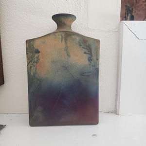 May include: A rectangular ceramic vase with a bottle-like neck. The vase has a gradient of colors, including brown, blue, and purple. The vase is displayed on a white shelf against a white wall.