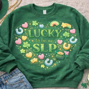 Op de afbeelding: Groene sweatshirt met een hartvormig ontwerp met de tekst "LUCKY to be an SLP", omringd door kleurrijke klavers, regenbogen, hoefijzers en sterren. De sweatshirt wordt gecombineerd met een spijkerbroek en accessoires met het thema St. Patrick's Day.
