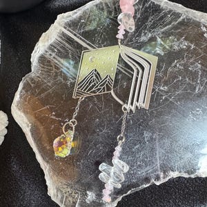 Könnte beinhalten: Silberfarbener Anhänger mit Berg- und Monddesign, mit einem schillernden Kristall und rosa und klaren Kristallakzenten. Der Anhänger hängt an einer Kette und ist wahrscheinlich ein dekorativer Artikel oder Schmuck.