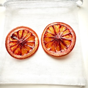 Pode incluir: Duas fatias de laranja sanguínea secas com tons vibrantes de vermelho e laranja, mostrando a intrincada estrutura interna da fruta. As fatias são apresentadas sobre um tecido branco texturizado, criando uma estética limpa e simples. Ideal para artesanato ou decoração.