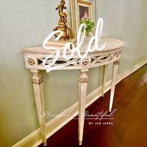May include: A cream-colored, half-moon console table with ornate carvings and gold accents. The table features a decorative openwork design along the apron and decorative detailing on the legs. A small statue and framed picture are on the table. The word "Sold" is written across the image.