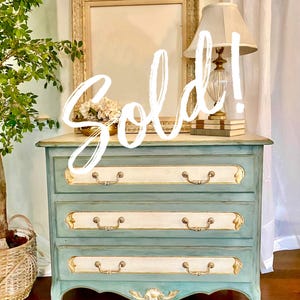 May include: A three-drawer chest of drawers painted in a distressed teal and cream color scheme. The drawers have gold accents and ornate handles. The word "Sold!" is written across the top. The bottom of the chest has a curved apron.