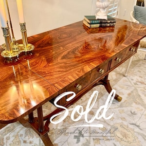 May include: A rectangular wooden desk with a high-gloss, swirling brown wood grain finish, featuring three front drawers with small brass knobs. The desk stands on four curved legs with brass casters and includes the text Sold Brushed to Beautiful BY JAN JARKE.