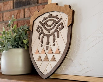 May include: A wooden wall hanging in the shape of a shield with a brown eye design and a pattern of triangles. The shield is on a wooden shelf with a potted plant.