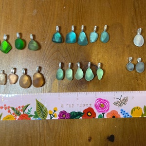 Op de afbeelding: Een verzameling van 20 zeeglas hangers met zilveren lussen. Het zeeglas is in verschillende tinten groen, blauw, wit en bruin. De hangers zijn gerangschikt in rijen op een houten oppervlak met een roze en bloemige liniaal.