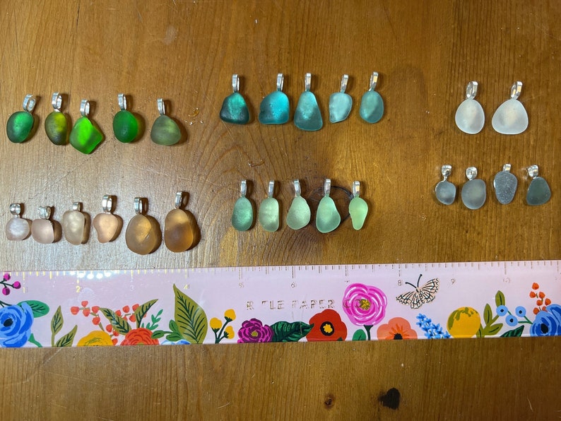 Op de afbeelding: Een verzameling van 20 zeeglas hangers met zilveren lussen. Het zeeglas is in verschillende tinten groen, blauw, wit en bruin. De hangers zijn gerangschikt in rijen op een houten oppervlak met een roze en bloemige liniaal.