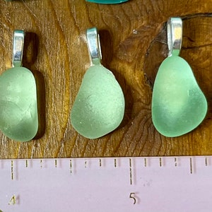 Op de afbeelding: Vijf zeeglas hangers met zilveren bedels. Het zeeglas is lichtgroen van kleur en heeft de vorm van een traan. De hangers zijn in een rij op een houten oppervlak gerangschikt met een liniaal eronder.