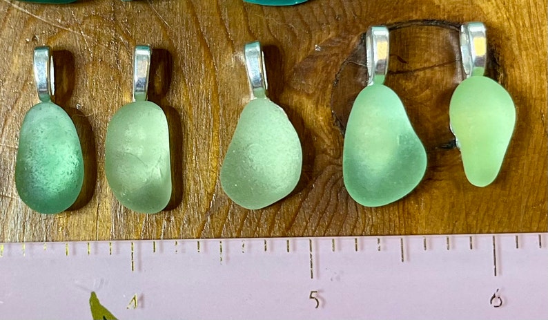 Op de afbeelding: Vijf zeeglas hangers met zilveren bedels. Het zeeglas is lichtgroen van kleur en heeft de vorm van een traan. De hangers zijn in een rij op een houten oppervlak gerangschikt met een liniaal eronder.
