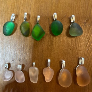 Op de afbeelding: Tien zeeglas hangers met zilveren bedels. Vijf hangers zijn groen en vijf zijn roze.