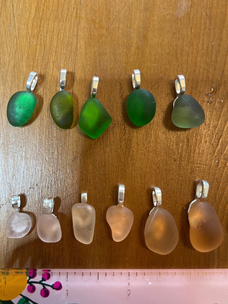 Op de afbeelding: Tien zeeglas hangers met zilveren bedels. Vijf hangers zijn groen en vijf zijn roze.