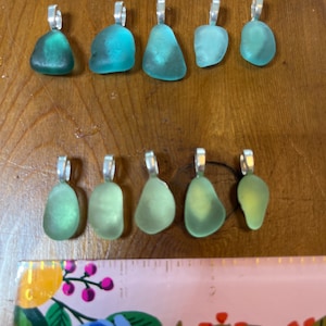Op de afbeelding: Tien hangers van zeeglas met zilveren springringen. Het zeeglas is in tinten groen en blauw.