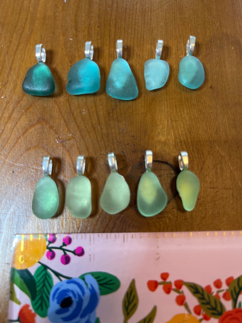 Op de afbeelding: Tien hangers van zeeglas met zilveren springringen. Het zeeglas is in tinten groen en blauw.