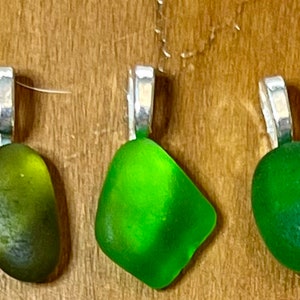 Op de afbeelding: Vijf groene zeeglas hangers met zilveren bedels. Het zeeglas is in verschillende tinten groen, van licht tot donker.
