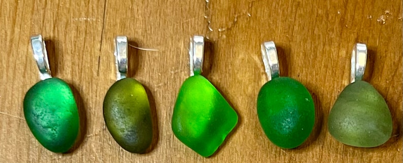 Op de afbeelding: Vijf groene zeeglas hangers met zilveren bedels. Het zeeglas is in verschillende tinten groen, van licht tot donker.