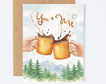 May include: Greeting card with a watercolor illustration of two orange mugs clinking, coffee splashing out. The text "You + Me" is above the mugs. A mountain scene and pine trees are in the background. The card is in a brown envelope.