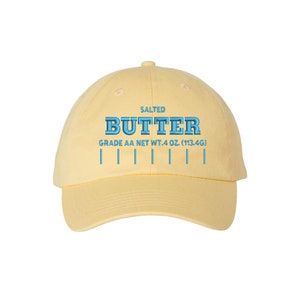 Puede incluir: Una gorra de béisbol amarilla con el texto "SALTED BUTTER GRADE AA NET WT. 4 OZ (113.4G)" bordado en azul.