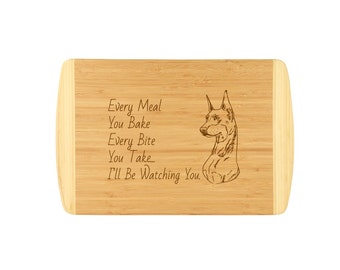 Könnte beinhalten: Ein braunes Bambus-Schneidebrett mit einem hellbraunen Holzrand. Das Brett ist mit einem gravierten Bild eines Dobermann-Hundes mit dem Text "Every Meal You Bake Every Bite You Take... I'll Be Watching You." versehen.