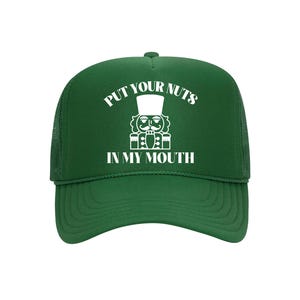 May include: A green trucker hat featuring a white nutcracker graphic and the text "PUT YOUR NUTS IN MY MOUTH". The hat has a mesh back and a curved peak.