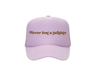 Könnte beinhalten: Helllila Trucker-Cap mit dem Text "Never lost a tailgate" in brauner Vintage-Schrift. Die Kappe hat einen gebogenen Schirm und eine Mesh-Rückseite.