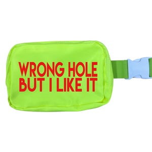 May include: A neon green fanny pack with the text "WRONG HOLE BUT I LIKE IT" in red. The pack has a green strap and a light blue buckle. The bag is rectangular and appears to be made of a nylon or similar material.