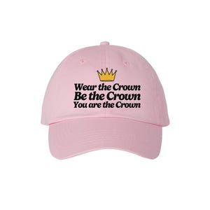 May include: A pale pink baseball cap with a curved peak. The cap displays the text "Wear the Crown, Be the Crown, You are the Crown" in black, with a gold crown graphic above the text.