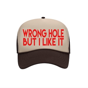 May include: A trucker hat with a tan front panel and a dark brown brim and mesh back. The hat features the red text "WRONG HOLE BUT I LIKE IT" across the front. The hat has a black button on top.