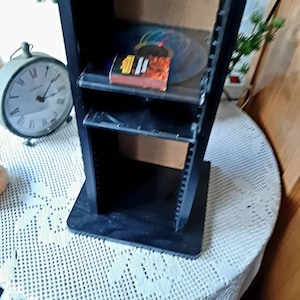 Könnte beinhalten: Schwarzer CD-Aufbewahrungsturm mit mehreren Regalen. Der Turm steht auf einer weißen Häkeltischdecke. Im Hintergrund ist eine Uhr zu sehen.