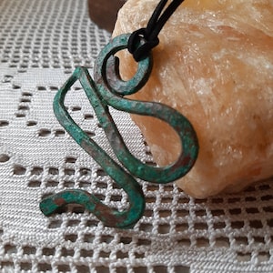 Pode incluir: Um pingente de metal verde e marrom em forma de cobra, pendurado em um cordão preto. O pingente está em uma superfície de crochê branca.