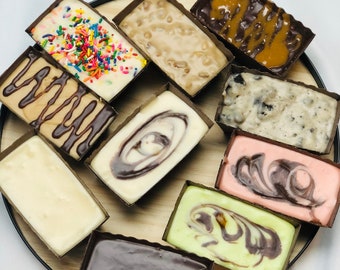 Puede incluir: Surtido de nueve cuadrados de fudge con diferentes sabores en recipientes de papel individuales sobre una bandeja de madera. Los sabores incluyen chocolate, vainilla, mantequilla de cacahuete y una variedad de sabores en espiral.