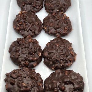 May include: Eight chocolate peanut butter patties on a white rectangular plate. The patties are round and have a smooth, glossy chocolate coating. The peanut butter is visible in the center of each patty.