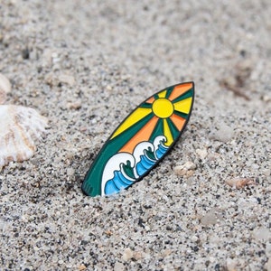 May include: A surfboard pin with a sun and wave design. The pin has a yellow sun, orange rays, green and yellow sections, and blue and white waves. The pin is set on sand with seashells.