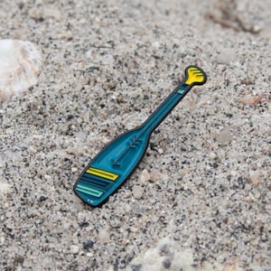 May include: A teal paddle-shaped enamel pin with yellow and black accents. The pin is lying on a sandy surface, suggesting a beach or nautical theme. The pin's design is a stylized representation of a paddle.