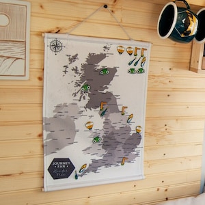 May include: A map of the British Isles with colorful pins marking locations. The map is gray and white, with a compass rose at the top. The text "JOURNEY * FAR * Wander Free" is at the bottom. Two mugs hang above the map.